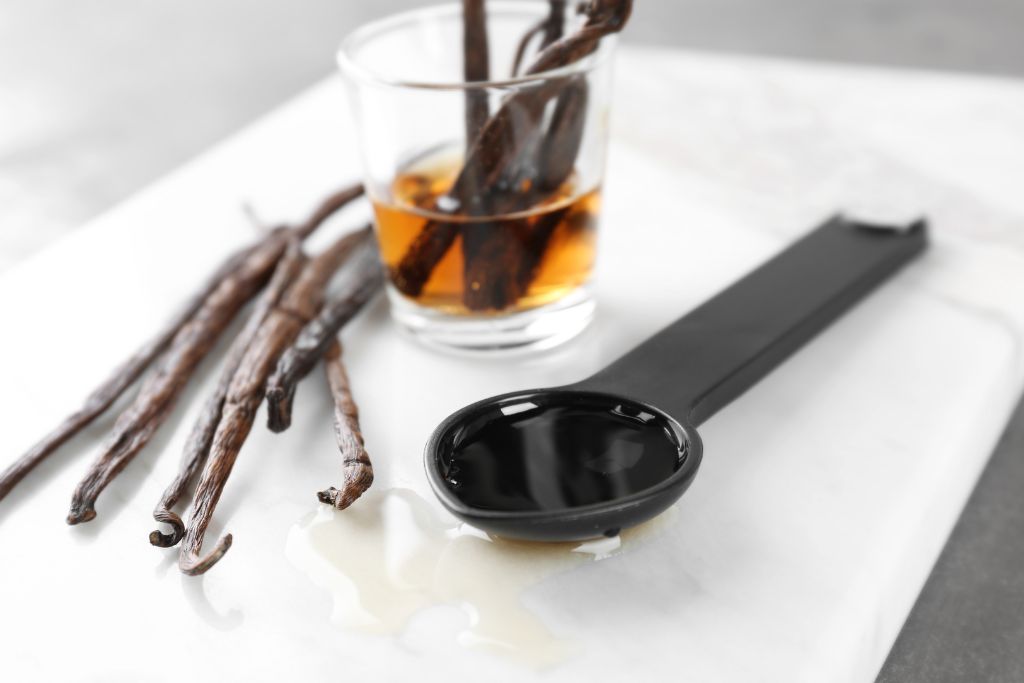 vanilla extract on a small shot glass together with vanilla dried sticks and spoon situated on white serving plate